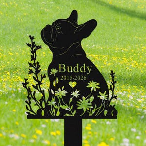 May include: Black metal garden stake featuring a silhouette of a French Bulldog. The dog's name, "Buddy", and dates "2015-2026" are displayed. The design includes floral and heart accents, with a stake for easy placement in a garden.