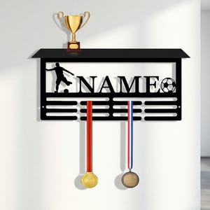 Może przedstawiać: Czarny metalowy wyświetlacz na trofea i medale piłkarskie. Wyświetlacz zawiera słowo "NAME" z piłką nożną i sylwetką piłkarza. Dwie medale wiszą poniżej. Złote trofeum na górze.
