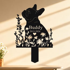 May include: Black metal garden stake featuring a French Bulldog silhouette, flowers, and foliage. The name "Buddy" and dates "2015-2026" are displayed. The stake has a T-shaped base for easy placement in a garden or yard.