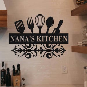 May include: Black metal kitchen sign with the text "NANA'S KITCHEN" and decorative scrollwork. The sign features silhouettes of kitchen utensils: a spatula, fork, whisk, spoon, and rolling pin.