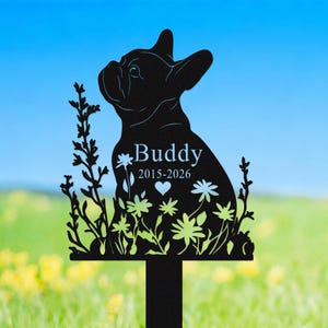 May include: Black metal garden stake featuring a silhouette of a French Bulldog. The stake includes the name "Buddy" with the years 2015-2026, a heart, and floral accents. The background is a blurred view of a garden.