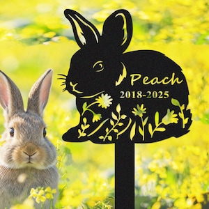 May include: A black metal garden stake featuring a rabbit silhouette with the name "Peach" and the years "2018-2025" in yellow. The stake is decorated with floral and leaf cutouts. A real rabbit is in the background.