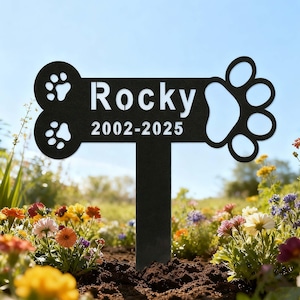 May include: A black metal pet memorial stake in a garden. The stake is shaped like a bone with a paw print design and the name "Rocky" and dates "2002-2025" in white lettering. The background features colorful flowers and a blue sky.