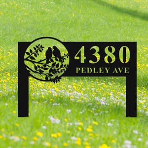 Könnte beinhalten: Schwarzes Metall-Adressschild mit der Nummer 4380 und dem Text "PEDLEY AVE". Das Schild zeigt ein kreisförmiges Design mit zwei Vögeln auf einem Ast. Das Schild ist auf zwei Pfosten montiert.