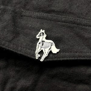 May include: A white enamel pin shaped like a horse with a visible black skeletal structure printed on its body. The pin is attached to a piece of black fabric. The horse is depicted in a running pose with its head turned slightly toward the viewer.