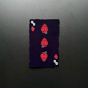 以下が含まれることがあります： 黒い背景と白い縁取りのあるトランプ。カードはハートの4で、黒の輪郭と中央に白い円を持つ赤いハートが4つあります。ハートはカードの中央に垂直に並んでいます。カードは少しすり切れ、しわがあります。