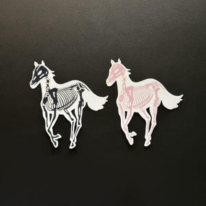 Peut inclure: Deux autocollants en forme de cheval. L'un est noir et blanc, représentant le squelette d'un cheval. L'autre est rose et blanc, montrant également un squelette de cheval. Les deux autocollants sont sur un fond noir.