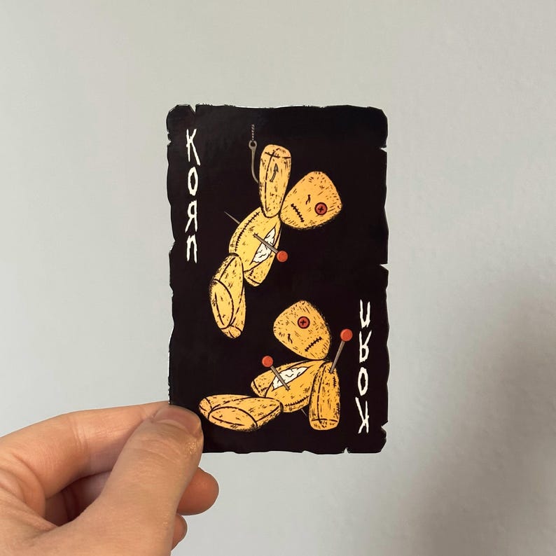 May include: A black card featuring two yellow voodoo dolls with red pins and stitched details. The word "Korn" is written vertically on both sides in white. The card is held by a hand.