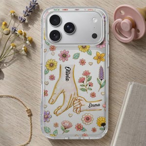 Könnte beinhalten: Transparente Handyhülle mit floralem Design und einer Grafik von zwei Händen, die sich halten. Die Namen "Olivia" und "Emma" sind auf der Hülle aufgedruckt. Die Hülle ist mit bunten Blumen verziert.