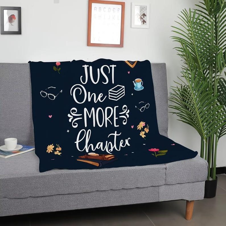 May include: A navy blue throw blanket with the text "JUST One MORE Chapter" in white script. The blanket features illustrations of books, glasses, flowers, and a teacup. The blanket is draped over a gray sofa.