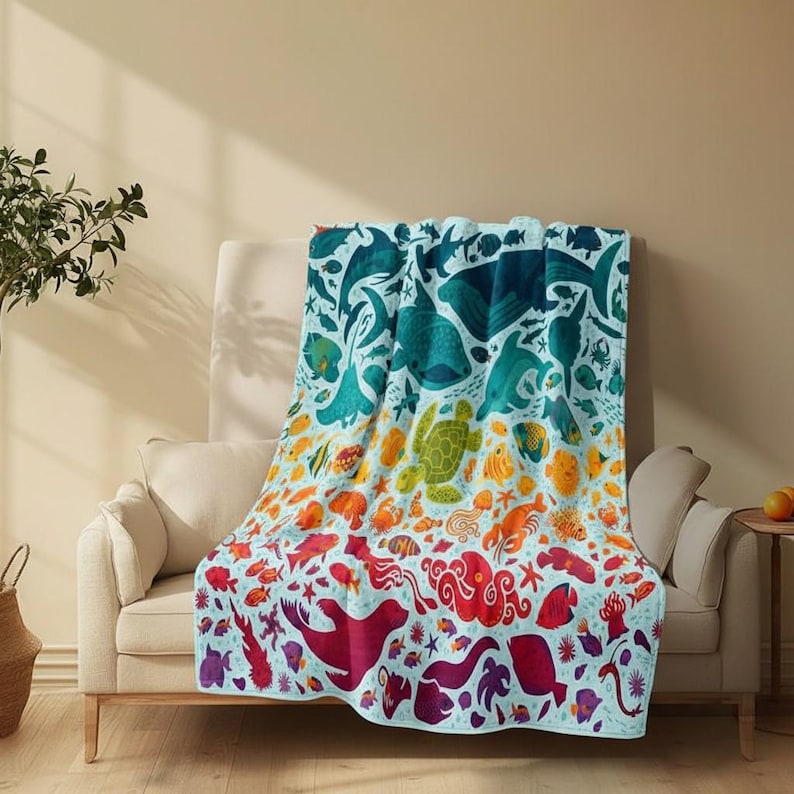 May include: A colorful throw blanket featuring an ocean-themed design. The blanket displays various marine life, including fish, turtles, and whales, in shades of blue, green, orange, and red. The blanket is draped over a beige sofa.