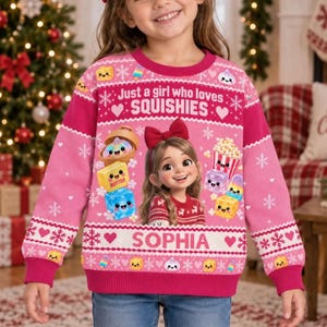 Könnte beinhalten: Ein rosafarbener Langarmpullover mit einem wiederkehrenden Schneeflocken- und Herzmuster, dem Text "Just a girl who loves SQUISHIES" und "SOPHIA" sowie einer zentralen Grafik einer Zeichentrickfigur mit roter Schleife und bunten Spielzeugen.