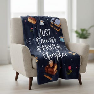 May include: A navy blue throw blanket draped over a beige armchair. The blanket features the text "JUST One MORE Chapter" in white script, along with illustrations of books, teacups, and flowers. The blanket is made of a soft material.
