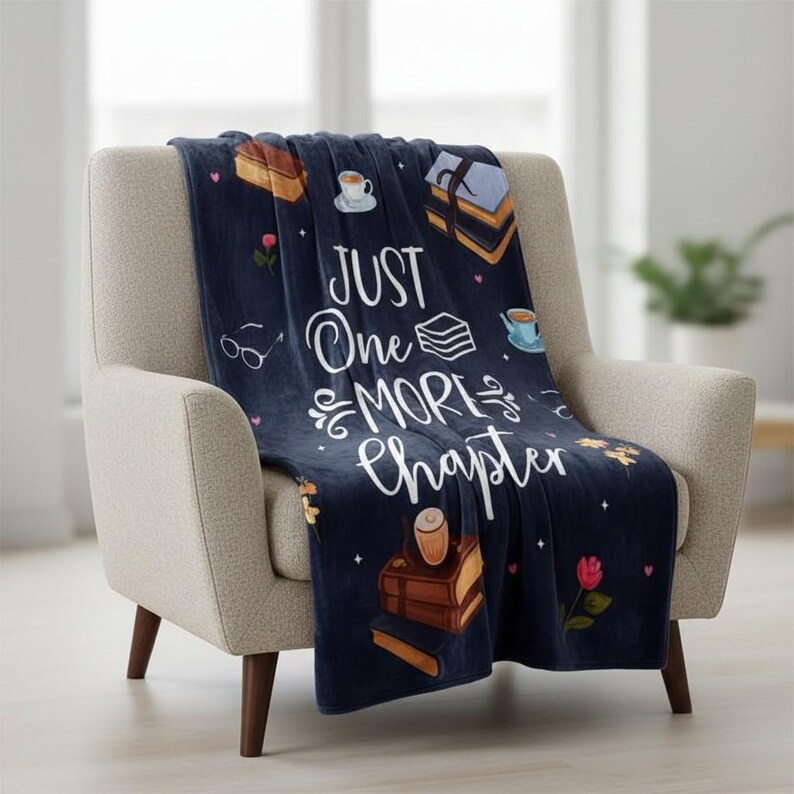May include: A navy blue throw blanket draped over a beige armchair. The blanket features the text "JUST One MORE Chapter" in white script, along with illustrations of books, teacups, and flowers. The blanket is made of a soft material.
