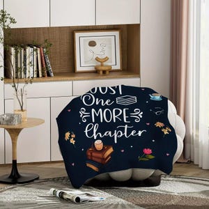 May include: A navy blue throw blanket with the text "Just One More Chapter" in white script. The blanket features illustrations of books, flowers, a teacup, and glasses. The blanket is draped over a gray armchair.