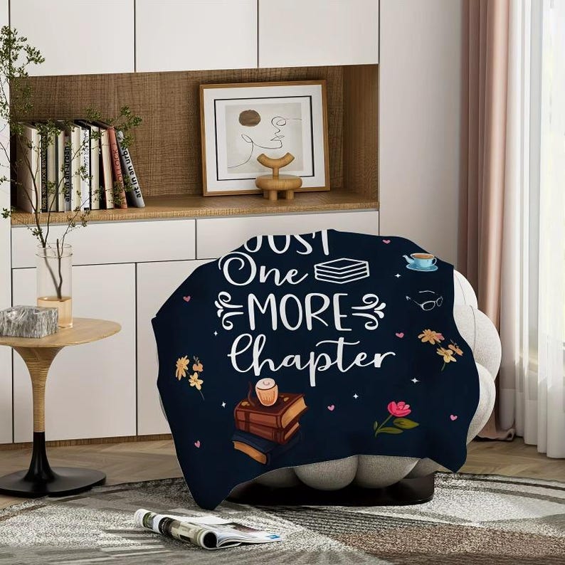 May include: A navy blue throw blanket with the text "Just One More Chapter" in white script. The blanket features illustrations of books, flowers, a teacup, and glasses. The blanket is draped over a gray armchair.