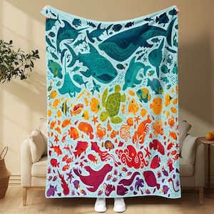 May include: A light blue blanket featuring a colorful illustration of various sea creatures. The design includes whales, sharks, fish, and other marine life in shades of blue, green, orange, and red. The blanket is draped over a sofa.