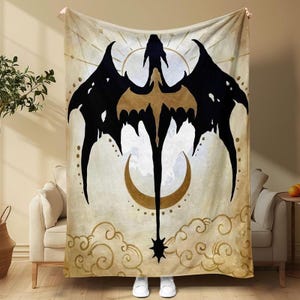 May include: A beige blanket featuring a black and gold dragon design. The dragon has large wings and a long tail, with a crescent moon and star at the bottom. The blanket is draped over a couch.