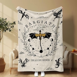 May include: Cream-colored blanket with a black and gold dragon design. The blanket features the text "BASGIATH WAR COLLEGE" and "DRAGON RIDER" with illustrations of dragons, stars, and celestial elements. The blanket is draped over a beige sofa.