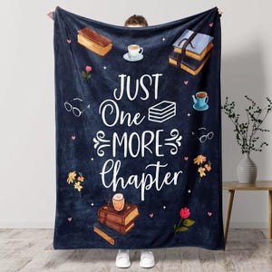 May include: A navy blue blanket with the text "JUST One MORE Chapter" in white script. The blanket features illustrations of books, teacups, eyeglasses, and flowers. The blanket is held up by a person wearing white shoes.