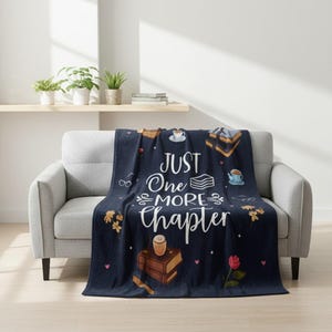 May include: A navy blue throw blanket draped over a gray sofa. The blanket features the text "JUST One MORE Chapter" in white script, along with illustrations of books, coffee cups, and flowers. The background is a solid navy blue.