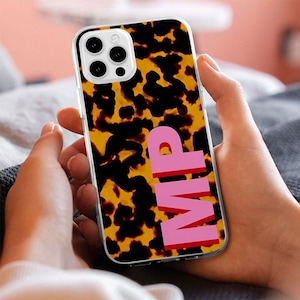 May include: A clear smartphone case featuring a brown and black tortoiseshell pattern with the capital letters MP printed in a bold, solid pink font vertically along the right side of the back panel, showcasing a triple camera lens cutout.