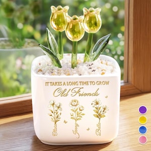 May include: A white ceramic planter with three glass tulip flowers. The planter has the text "IT TAKES A LONG TIME TO GROW Old Friends" and floral designs with names. The tulips are yellow and green.