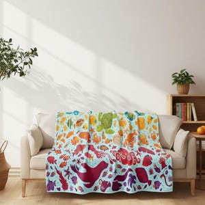 May include: A colorful blanket featuring a vibrant array of sea creatures, including fish, turtles, and octopuses. The design transitions from blue to red, creating a gradient effect. The blanket is draped over a beige sofa.