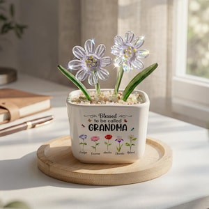 May include: A white ceramic flower pot with the text "Blessed to be called GRANDMA" and flower illustrations. The pot holds two clear, decorative flower ornaments with green stems and leaves, resting on a wooden tray.