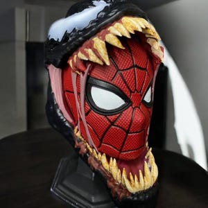 Puede incluir: Un busto de cabeza de Spider-Man rojo y negro con un busto de cabeza de Venom negro y blanco mordi&eacute;ndolo. El busto de cabeza de Venom tiene dientes afilados y una lengua larga.