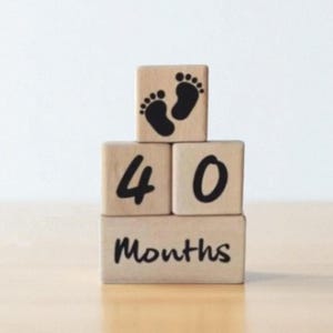 May include: Wooden blocks stacked to read "40 Months" with a pair of black baby footprints on top. The blocks are light brown and sit on a light brown surface, with a white background.