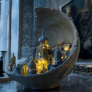 May include: A gray, crescent-shaped decorative piece featuring a miniature city with illuminated windows and towers. The city is nestled within the crescent, creating a unique and artistic display. The piece is likely made of a stone-like material.