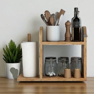 Puede incluir: Estantería de cocina de madera de dos niveles con varios elementos. Incluye un portarrollos de papel, una pequeña planta en maceta, tarros, una botella y utensilios. La estantería es de madera clara.