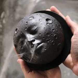 May include: A black ceramic incense burner shaped like the moon with a face and craters. Smoke is rising from the burner.