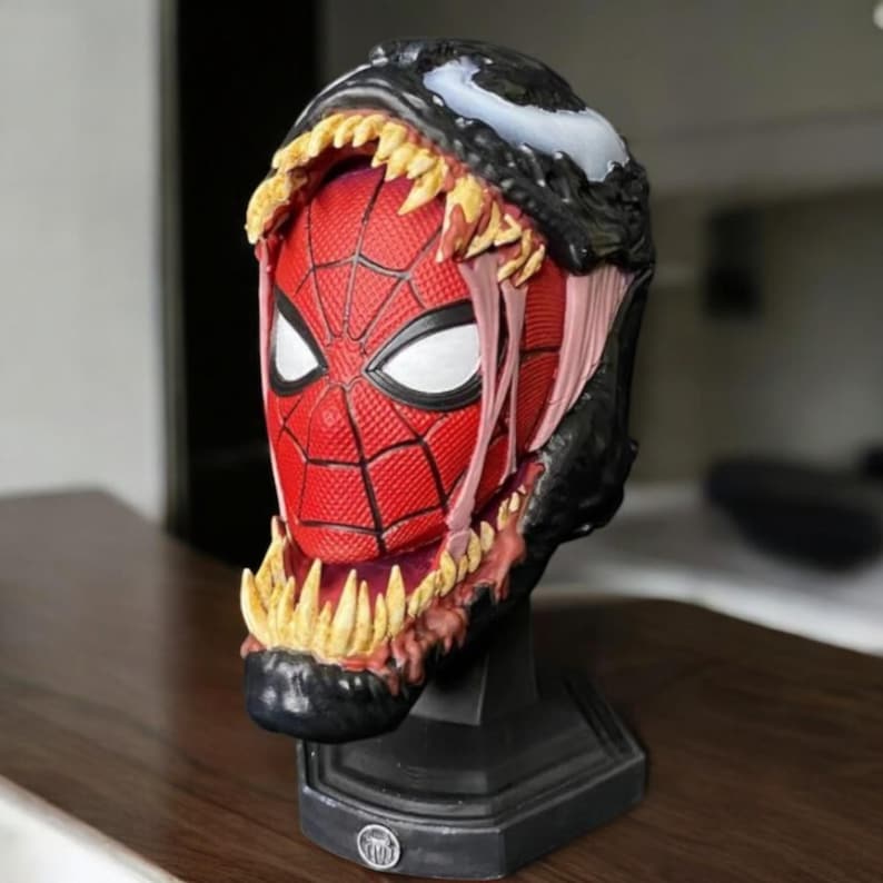 Puede incluir: Un busto negro y rojo de Spider-Man con un simbionte Venom negro y blanco que cubre parcialmente su cabeza. El simbionte Venom tiene dientes afilados y una lengua blanca. El busto est&aacute; sobre una base negra.