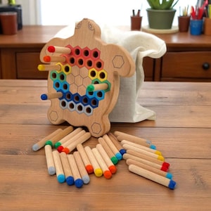 Può includere: Gioco puzzle a forma di tartaruga in legno con pioli colorati. Il gioco presenta una base in legno marrone chiaro con fori esagonali e pioli in vari colori, tra cui rosso, arancione, giallo, verde, blu e grigio. I pioli sono sparsi.