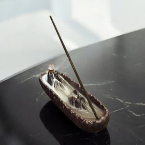May include: A brown ceramic incense burner shaped like a mountain range with a single incense stick burning. The incense smoke is rising from the burner and creating a wispy cloud.