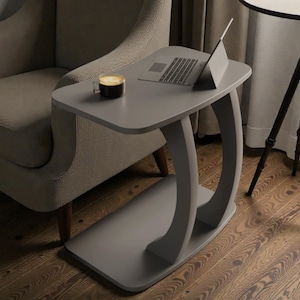 May include: A gray, modern side table with a curved base and a rectangular top. A laptop and a candle sit on the table. The table is next to a beige armchair and a tripod table.