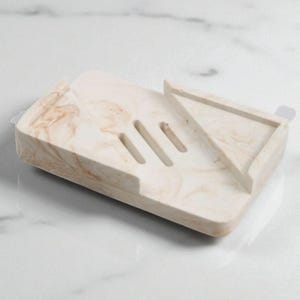 Puede incluir: Un jabonera rectangular con un patrón veteado beige y marrón. La jabonera tiene tres ranuras para el drenaje y una sección triangular elevada. La jabonera está hecha de un material sólido.