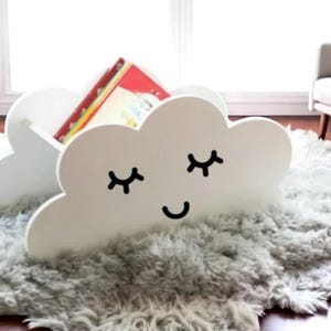 Pode incluir: Uma estante branca em forma de nuvem com um rosto sorridente e olhos fechados, segurando livros coloridos. A estante está sobre um tapete cinza fofo, ideal para um quarto infantil.
