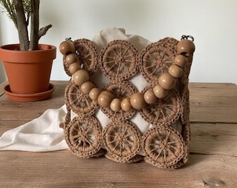 May include: A handcrafted, square-shaped handbag with a unique design. The bag features a woven, wheel-like pattern in a light brown color. It has a wooden beaded handle and a cream-colored interior. The bag is placed on a wooden surface.