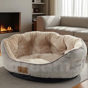 May include: A round pet bed with a plush, beige interior and a gray exterior. The bed has a low profile and a black base. A small brown label with text is visible on the side. The bed is on a gray rug.