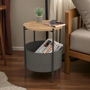May include: A round side table with a light wood top and a dark gray metal frame. The table has a built-in storage basket and holds a newspaper, a small plant, a remote control, and a book. The table is next to a wooden chair.