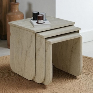 May include: A set of three nesting tables in a light beige stone finish. The tables have a square top and rounded edges. Small decorative items sit on the top table. The tables are on a round, woven jute rug.