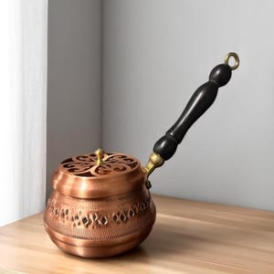May include: A copper-colored, decorative pot with a lid and a long, black wooden handle. The pot has an ornate design with cut-out patterns and a gold-colored accent. The handle is attached with a gold-colored fitting.