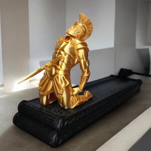 Puede incluir: Una estatua dorada de un soldado romano arrodillado sobre una base negra. El soldado lleva armadura y casco. La estatua es una pieza decorativa.