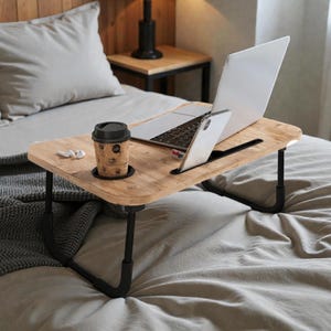 Puede incluir: Un escritorio de madera para portátil con patas negras está sobre una cama. Una computadora portátil, un teléfono inteligente y una taza de café están en el escritorio. El escritorio tiene una ranura para el teléfono y un portavasos. La cama tiene ropa de cama gris y una manta gris.