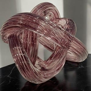 Może przedstawiać: Duża, abstrakcyjna rzeźba szklana w ciemnoróżowym kolorze. Szkło jest skręcone i splecione w złożony kształt, tworząc unikalny i nowoczesny design.