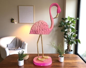 May include: A large, pink flamingo sculpture with gold-colored legs and beak. The flamingo stands on a round, pink base. The sculpture is a decorative item, likely for a home or garden. The flamingo's body has a textured surface.