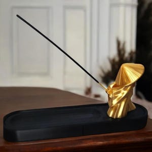 May include: Gold incense holder shaped like a person wearing a conical hat, sitting on a black rectangular base. A black incense stick is placed in the holder.